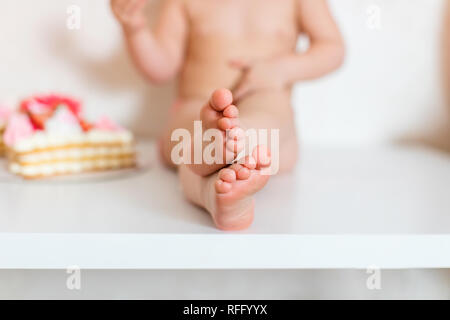 Little blonde baby girl two years old in pink pants sitting on the white table near her birthday cake and different pink sweets on the table. Stock Photo