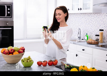 Smiling Young Woman Using Cellphone Near Fresh Fruits And Vegetables On Kitchen Counter Stock Photo
