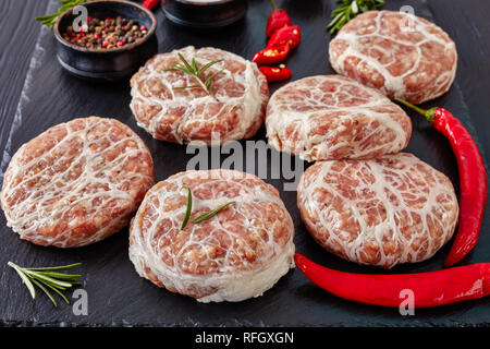 meat delicatessen - raw Atriaux of ground pork wrapped in caul fat, swiss cuisine, type of bratwurst on a slate tray with rosemary and salt, on a wood Stock Photo
