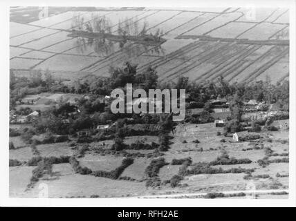 Black and white photograph, showing an aerial view or birds-eye view of a Vietnamese hamlet or small village, with flooded farmland (likely rice paddies) visible in the background, photographed during the Vietnam War, 1968. ()