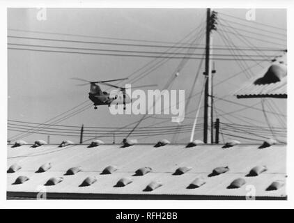 Black and white photograph, showing a tandem rotor helicopter, likely a Boeing CH-47 Chinook, in flight, with electrical wires and the roof of a barrack or administrative building visible in the foreground, photographed during the Vietnam War, 1968. ()
