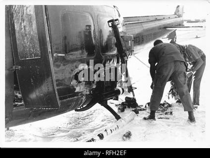 Black and white photograph, showing two men in military uniforms, bending over to survey the damage sustained by a Bell UH-1 Iroquois ('Huey') helicopter, with a light colored substance (possibly sand, snow, or foam) visible on the ground, photographed during the Vietnam War, 1968. ()
