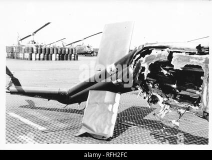 Black and white photograph, showing the heavily damaged tail boom and engine mount section of a military helicopter, likely a Bell UH-1 Iroquois ('Huey') with barrels and more helicopters visible in the background, photographed during the Vietnam War, 1968. () Stock Photo