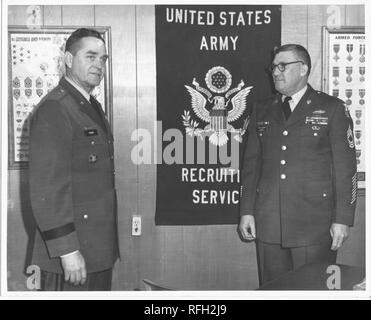 Black and white photograph of two middle-aged men, each standing in three-quarter length view, wearing US Military uniforms, with Brigadier General Frank L Gunn (left) shown in partial profile, with a relaxed expression on his face, the man at right wears glasses and faces General Gunn, posters with images of insignias, badges, and medals, and a sign with the text 'United States Army Recruitment Services, ' are visible in the background, photographed during the Vietnam War, 1967. () Stock Photo