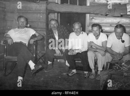 Black and white photograph of five unshaven, middle-aged men, sitting together facing the camera; one sits in a rocking chair smoking a pipe, and the other four (three smoking pipes) sit closely next to each other on the bottom bunk of a camp bed, photographed during a hunting and fishing trip located in Alaska, USA, 1955. () Stock Photo