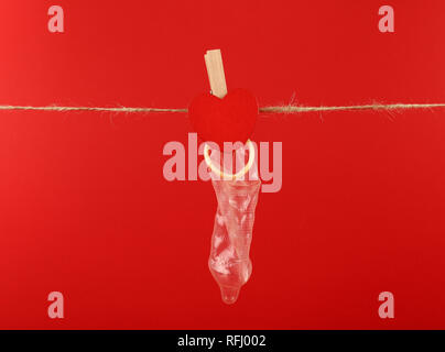 Close up one condom hanging on twine washing line with wooden clothespin with heart shaped sign over red background with copy space, low angle side vi Stock Photo