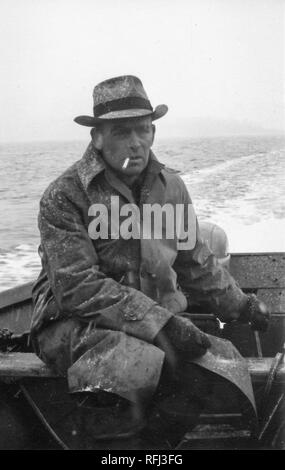 Black and white photograph of a middle-aged man, with a cigarette between his lips, wearing a snow-dusted coat, gloves, and hat, sitting in the stern of an open dinghy, steering the tiller of an outboard motor with one hand, with a light wake and a shadowy outline of land visible in the background, photographed during a hunting and fishing trip located in Alaska, 1955. () Stock Photo