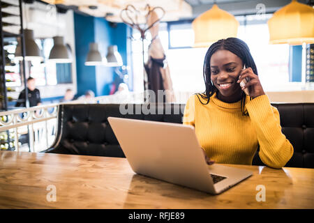 Closeup portrait of young african woman relaxing in cafe with laptop and making phone call Stock Photo
