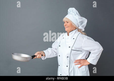 Senior woman chef studio standing isolated on gray with frying pan cheerful side view Stock Photo