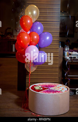 Colorful macarons cake . Paper Box with french dessert - macaroons on gray concrete table with festive decoration. Red and purple flowers . Red Balloo Stock Photo