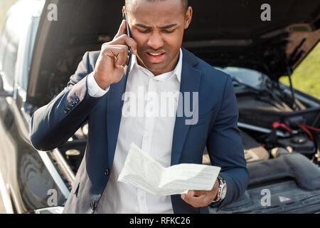 Young man leaning on car with opened hood holding insurance calling on smartphone frowning close-up Stock Photo