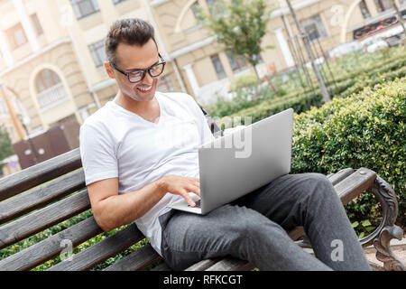 Freelance job. Young man on city street sitting on bench working on laptop smiling cheerful Stock Photo