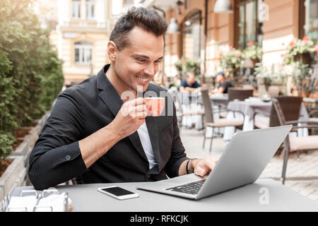 Lunch time. Young man in suit sitting at cafe on city street drinking hot espresso browsing internet on laptop smiling cheerful Stock Photo