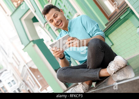 Outdoors leisure. Young man sitting on city street playing game on digital tablet smiling excited Stock Photo