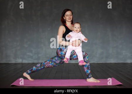 Beautiful sporty fit woman practices yoga asana Virabhadrasana holding her baby. Studio shoot Stock Photo