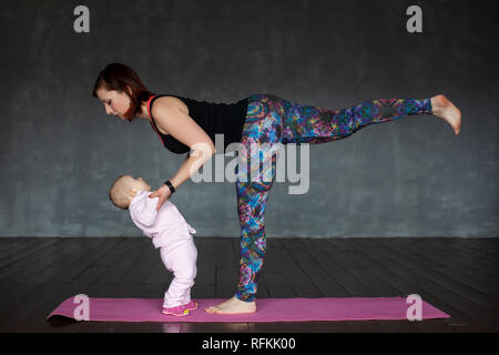 Beautiful sporty fit woman practices yoga asana Virabhadrasana holding her baby. Studio shoot Stock Photo