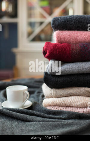 Pile of colorful sweaters with hot drink white cup on the side, background with copy space, cozy hygge wallpaper. Stock Photo