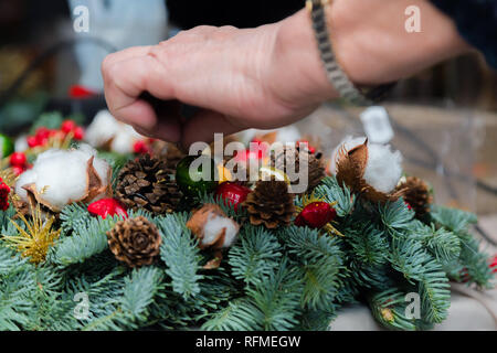 Christmas wreath weaving workshop. Woman hands decorating holiday wreath made of spruce branches, cones and various organic decorations on the table Stock Photo