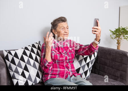 A very old senior Caucasian grandmother with gray hair and deep wrinkles sits home on couch in jeans and red checkered shirt and uses her smart phone  Stock Photo
