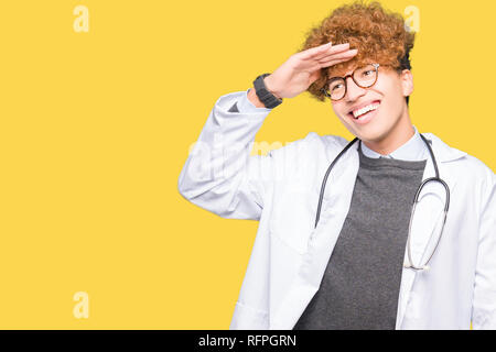 Young handsome doctor man wearing medical coat very happy and smiling looking far away with hand over head. Searching concept. Stock Photo