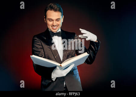 Professional Occupation. Magician in suit and gloves standing isolated on wall making spell with magic book smiling excited Stock Photo