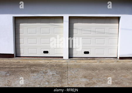 Two modern white rollup garage doors with cracked concrete driveway mounted on white wall on warm sunny day Stock Photo