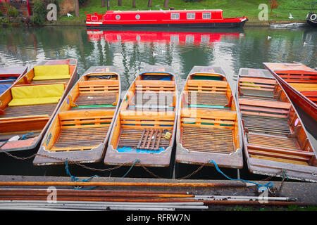 Collection of punts moored side by side at a bank of the river Cam opposite a red narrow boat reflected in the water on the opposite shore. Stock Photo