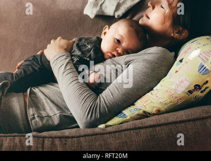 Mother taking rest sleeping on a couch with her baby on her chest. Close up of a woman lying on a sofa with her baby resting on her. Stock Photo