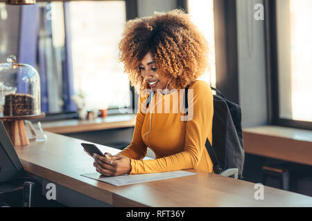 Smiling afro american female wearing backpack standing at the billing counter looking at her mobile phone. Woman making payment using her mobile phone Stock Photo