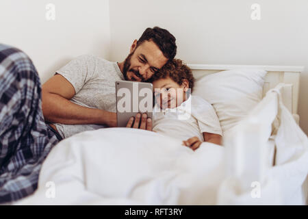 Father and son lying on bed watching a movie on tablet pc. Man lying on bed with his kid holding a tablet pc in hand. Stock Photo