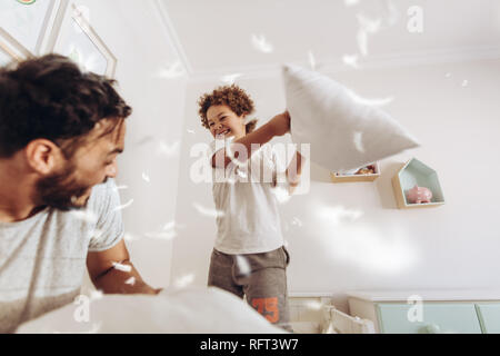Happy father and son having a pillow fight on bed with feathers flying around. Father and son having fun playing at home. Stock Photo