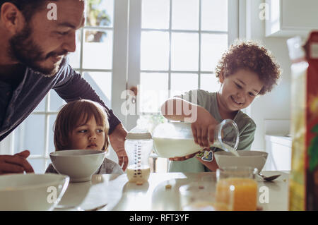 Man watching his kid pour milk in his breakfast bowl. Father and kids sitting at the table preparing breakfast. Stock Photo