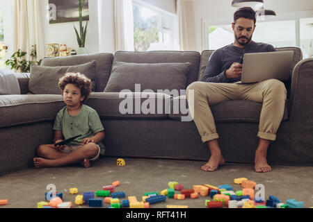 Man working on laptop from home sitting on couch. Boy sitting on floor with building blocks in front and his father working on laptop. Stock Photo