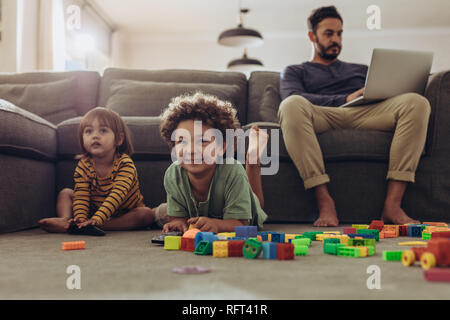 Smiling kids playing with building blocks and watching television at home. Man babysitting his kids and working from home. Stock Photo