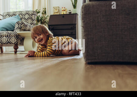 Smiling kid sitting on floor at home. Cheerful kid hiding behind a couch playing hide and seek. Stock Photo