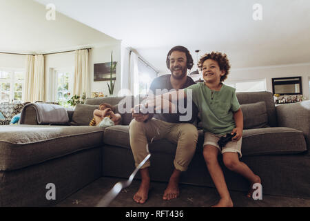 Cheerful father and son enjoying video game at home. Man playing video game sitting on couch at home with his son. Stock Photo