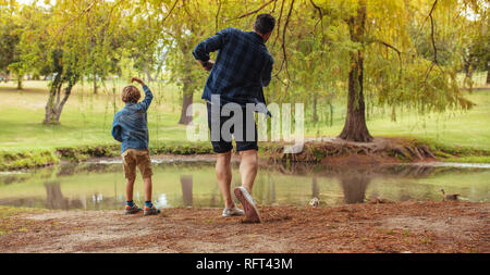 Rear view of father and son throwing rocks in pond at park. Man and little boy having fun at the park. Kid with father throwing pebbles in pond. Stock Photo