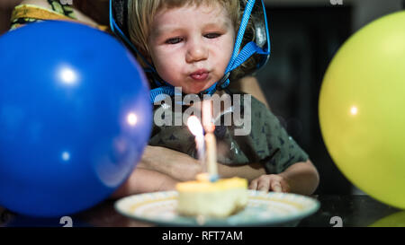A portrait of a happy birthday boy wearing a blue helmet and blowing off the birthday candles. Blue and yellow air balloons in the foreground. Stock Photo