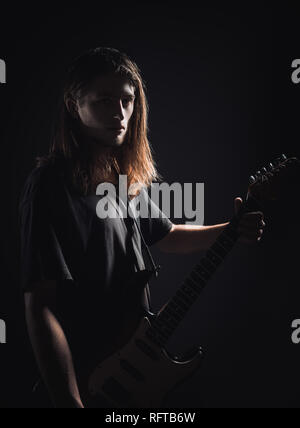 Dramatic studio portrait: a handsome long-haired young man (rock musician) holds electric guitar in hands Stock Photo