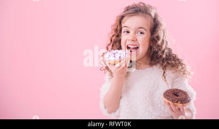 Portrait of a little joyful cute curly girl who eats donuts, on a pink background, a place for text Stock Photo
