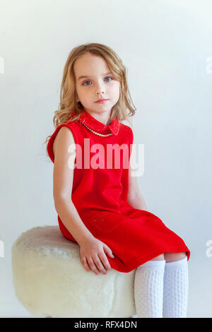 Sweet happy little girl in red dress sitting on chair against white wall background in bright room Stock Photo