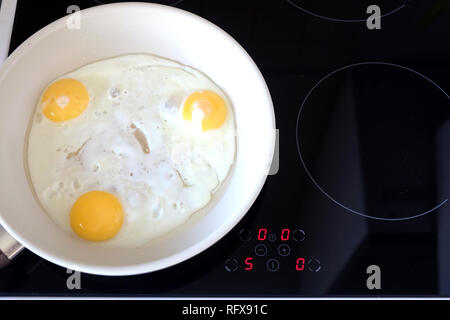 Ready breakfast on stove. Three appetizing fried eggs in white pan on modern kitchen induction stove with electronic control panel top view closeup Stock Photo