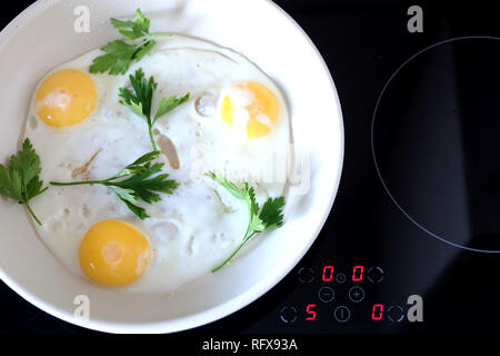 Ready breakfast on stove. Three appetizing fried eggs with parsley in white pan on modern kitchen electric stove with electronic control panel top vie Stock Photo