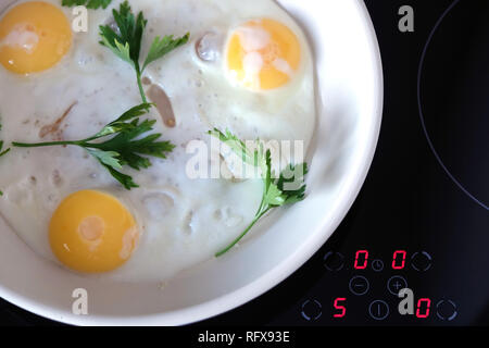 Ready breakfast on stove. Three appetizing fried eggs with parsley in white pan on modern kitchen electric stove with electronic control panel top vie Stock Photo