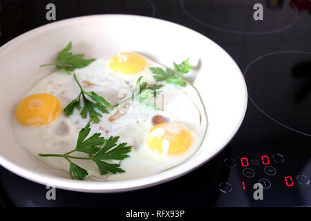 Ready breakfast on stove. Three appetizing fried eggs with parsley in white pan on modern kitchen electric stove with electronic control panel top vie Stock Photo