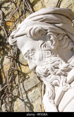 Close up of profile of head of stone statue of an old man with a long curly beard and a hat. Outdoors on a very sunny day. Stock Photo