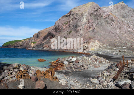 Remnants of machinery and equipment and a ships anchor lie abandoned on the rocky beach. Stock Photo