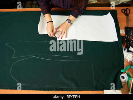 Tailor worktable in the studio. Close-up of woman hands making chalk markings of paper patterns on green fabric copy space surface before cutting deta Stock Photo