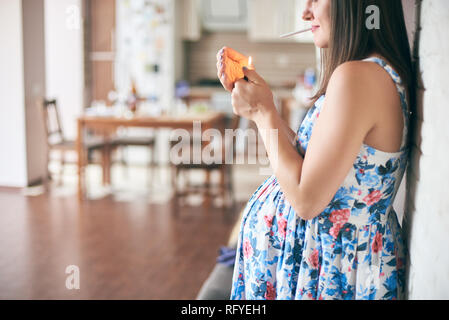 Side view of pregnant woman in dress standing in kitchen and keeping in hands cigarette and lighter. Future mother smoking and leading unhealthy lifes Stock Photo