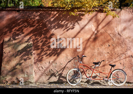 Modern comfortable orange tandem double bicycle with white wheels on paved empty sidewalk leaned against old plastered cracked red wall on bright sunn Stock Photo